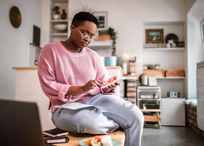A young woman in a pink sweater sits on her coffee table with a piece of paper resting on her thigh while she looks intently at her phone. 