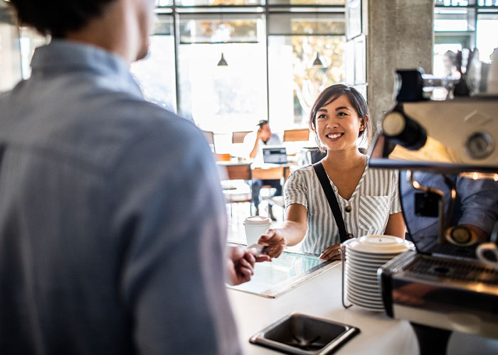 A woman in a white shirt white black stripes smiles and hands her credit card to the cashier at a coffee shop. 
