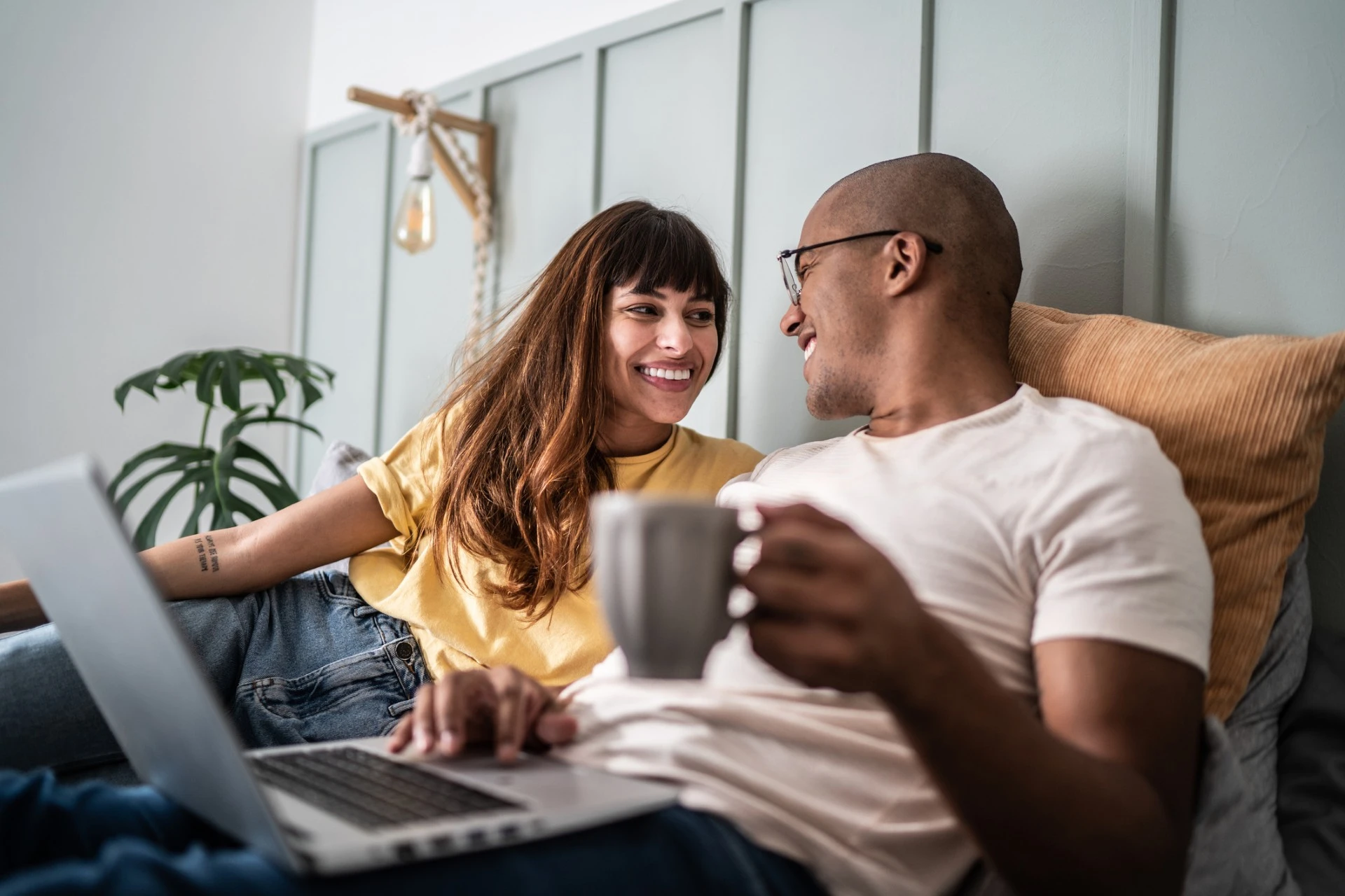 A man and women sit together and smile at each other while using a laptop.