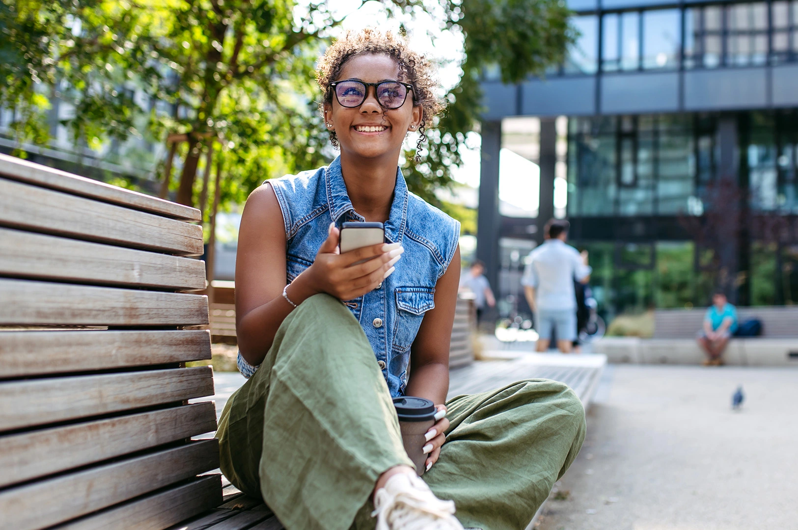 A young woman sits on a park bench smiling while holding a smartphone. 