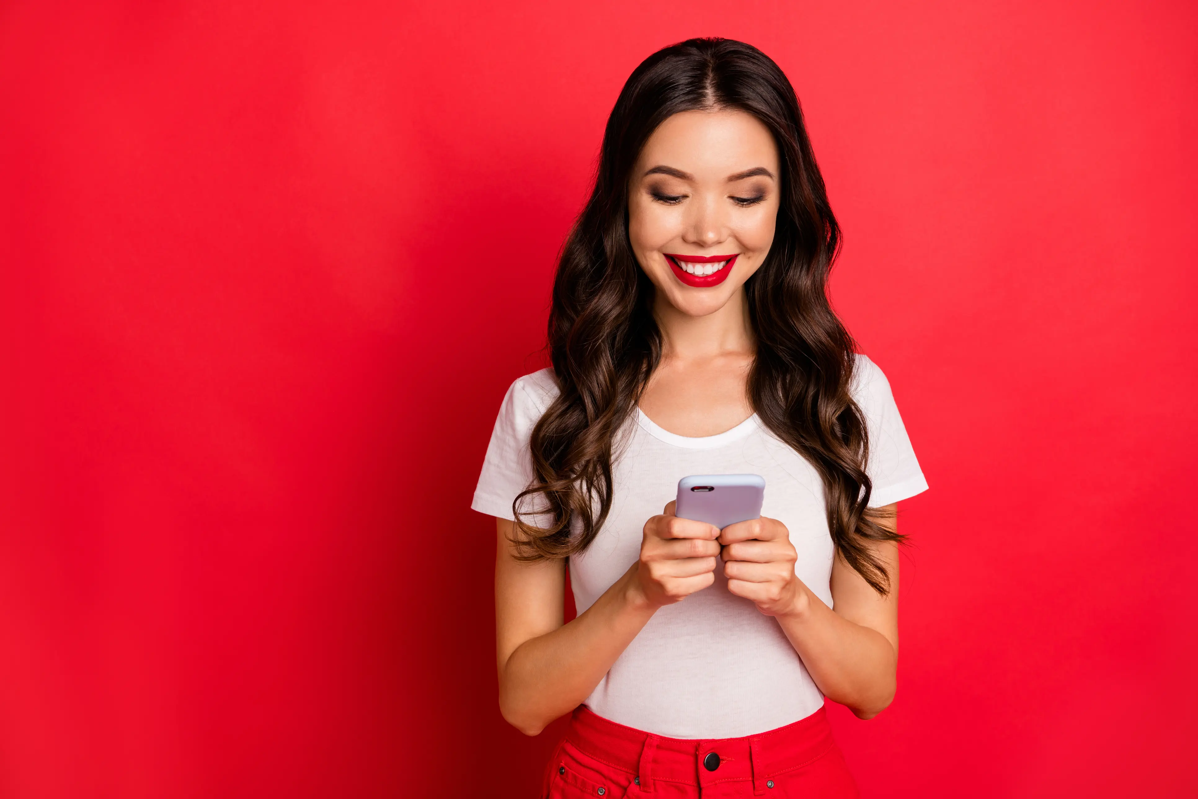 A woman in a white t-shirt stands in front of a red background looking down and smiling at her smartphone that she holds in both hands. 