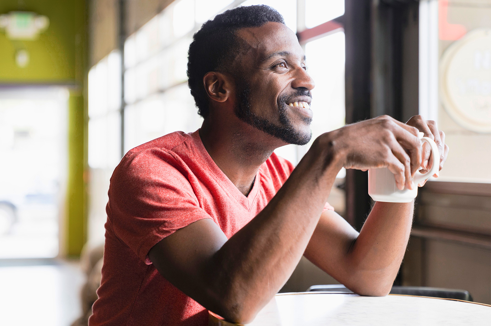 A smiling man in an orange t-shirt sits at a cafe table holding a cup of coffee.