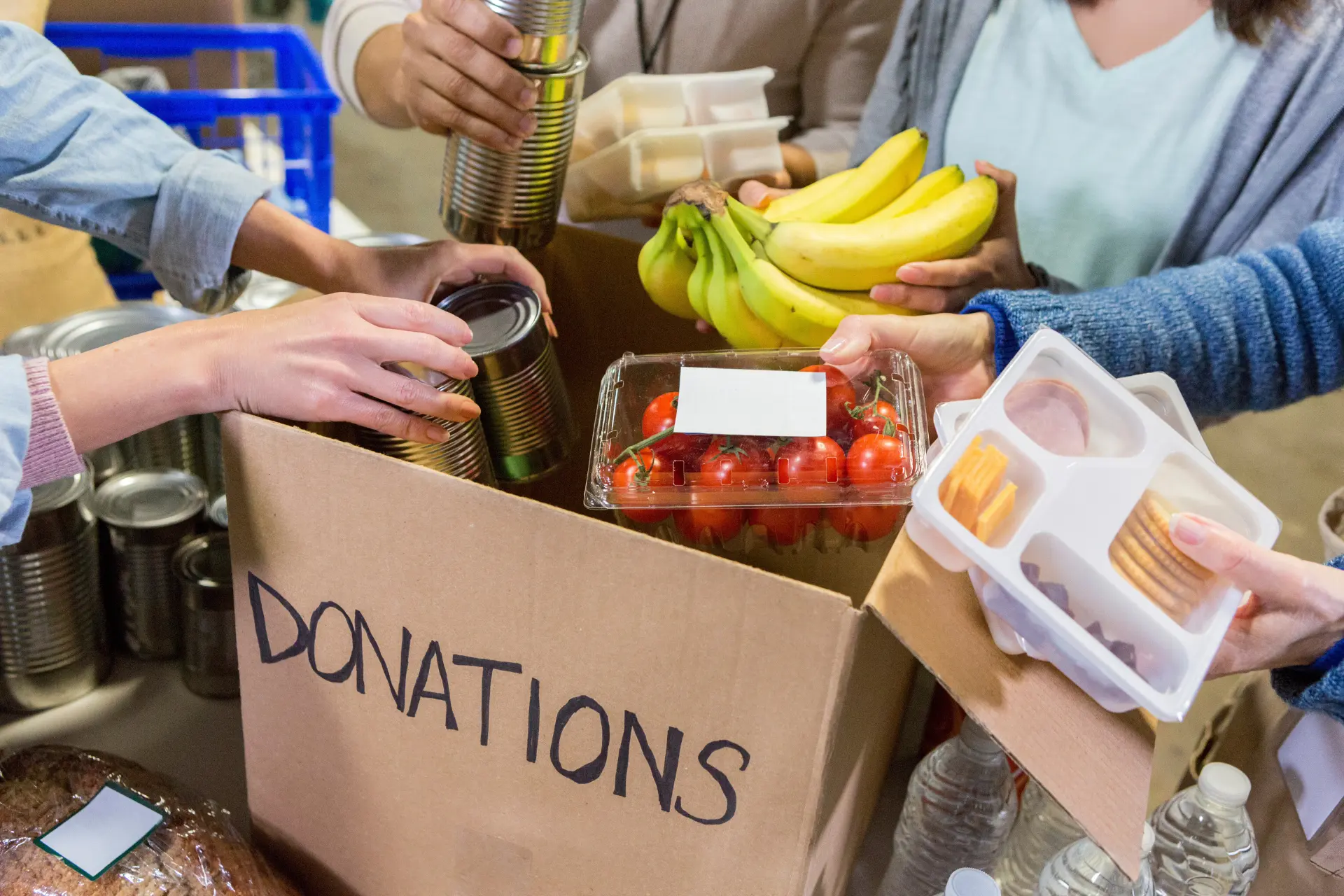 A cardboard box labelled "Donations" is being filled with foods like bananas and tomatoes by a group of people.