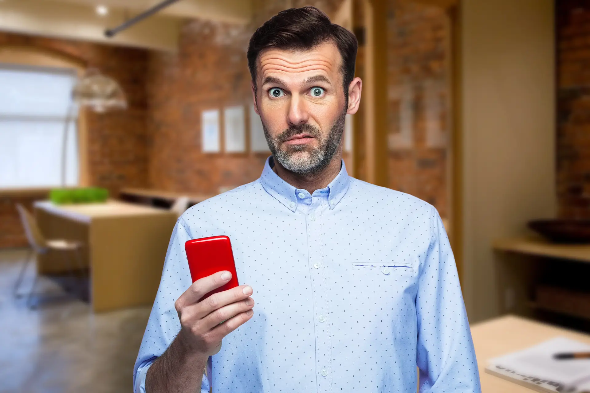A man in a blue buttoned shirt holding a red smartphone faces the camera with a concerned look on his face. 