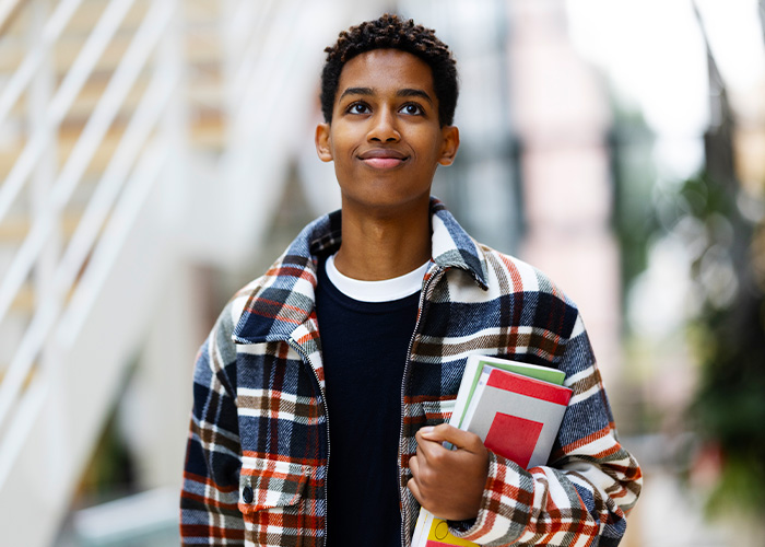 A young man in a plaid jacket smiles and looks up hopefully as he carries textbooks by his side.