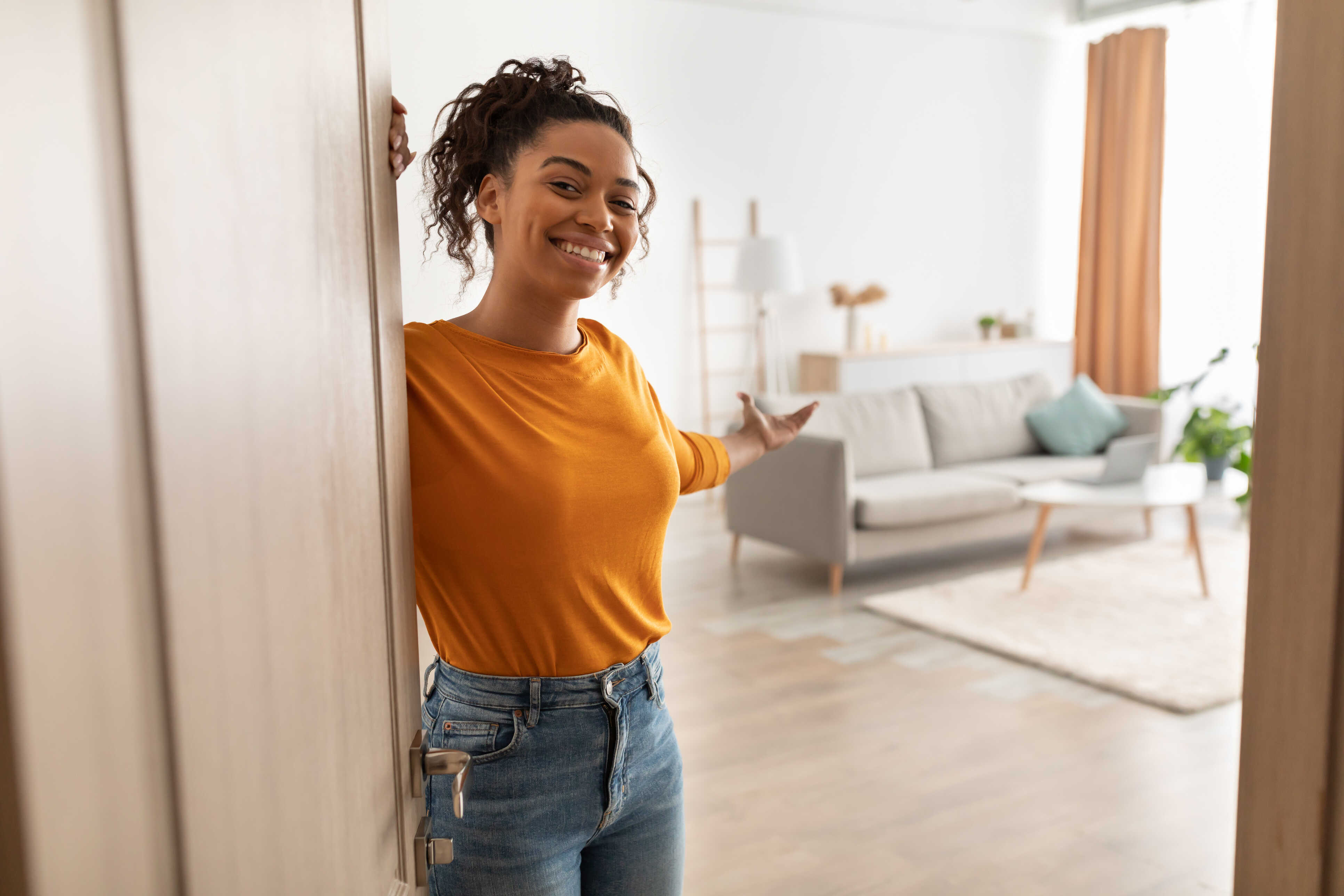 A smiling women in an orange shirt extends her arm to welcome a guest into her new home.