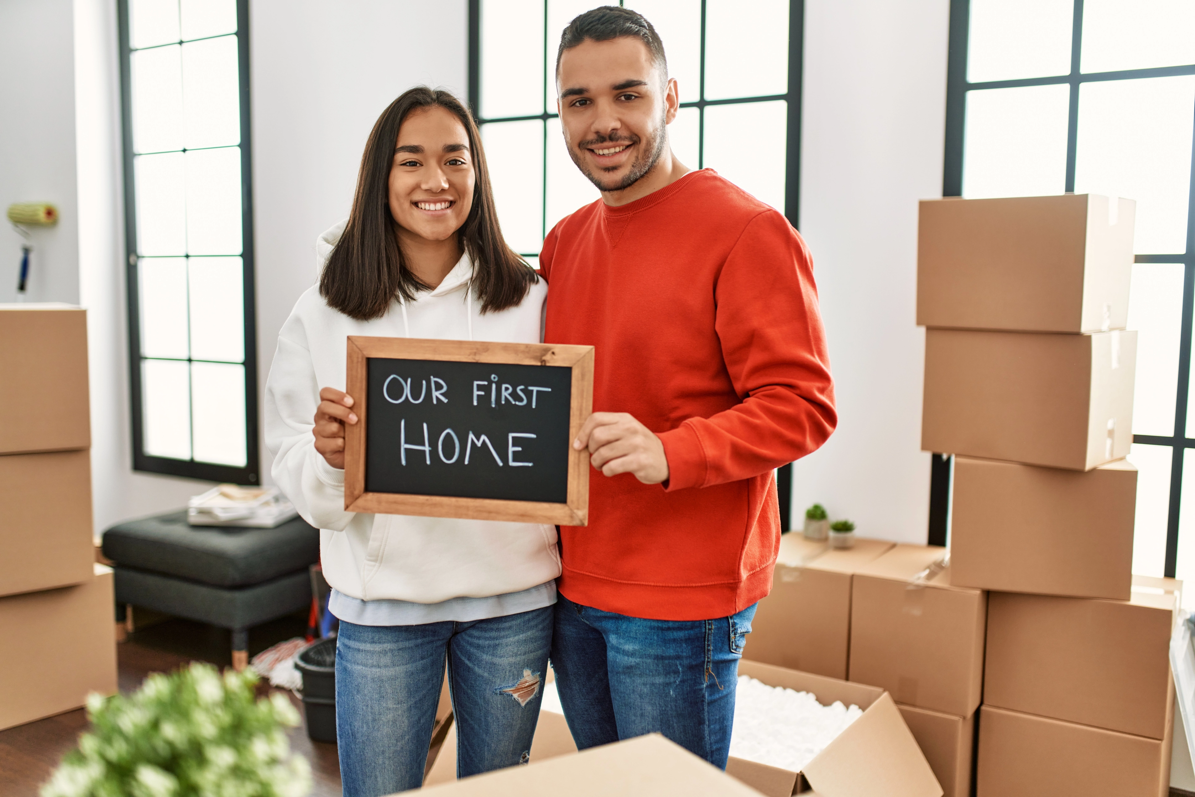 A young couple holds up a sign together saying "Our First Home".